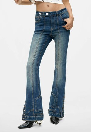 Jeans de mezclilla azul con corte acampanado, detalles en la costura frontal y dobladillos con botones, combinados con botas negras puntiagudas y un top corto.