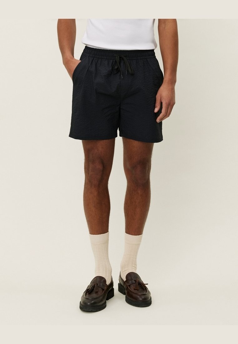 Homme portant un short noir à cordon, des chaussettes blanches et des mocassins à glands marron foncé, debout les mains dans les poches devant un fond uni.