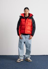 Rød og sort puffer vest med en tætsiddende design lagdelt over en sort skjorte, parret med løse blå jeans og sorte og hvide sneakers.