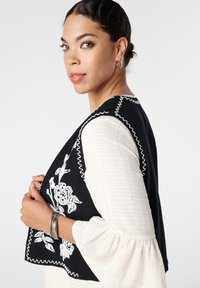 Zwarte geborduurde vest met witte bloemmotieven, gegolfde randen, gedragen over een textuur wit langhangend shirt. Zilveren armbanden om de pols.