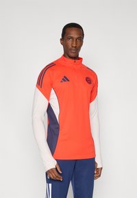 Rød atletisk topp med halv glidelås med hvite og marineblå detaljer, med tre striper på skuldrene og en rund laglogo på brystet.