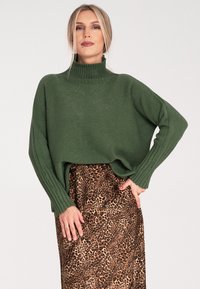Zielony sweter robiony na drutach z wysokim kołnierzem i ribbowymi rękawami, w połączeniu z midi spódnicą w leopardzi wzór. Stylizacja charakteryzuje się luźnym krojem.