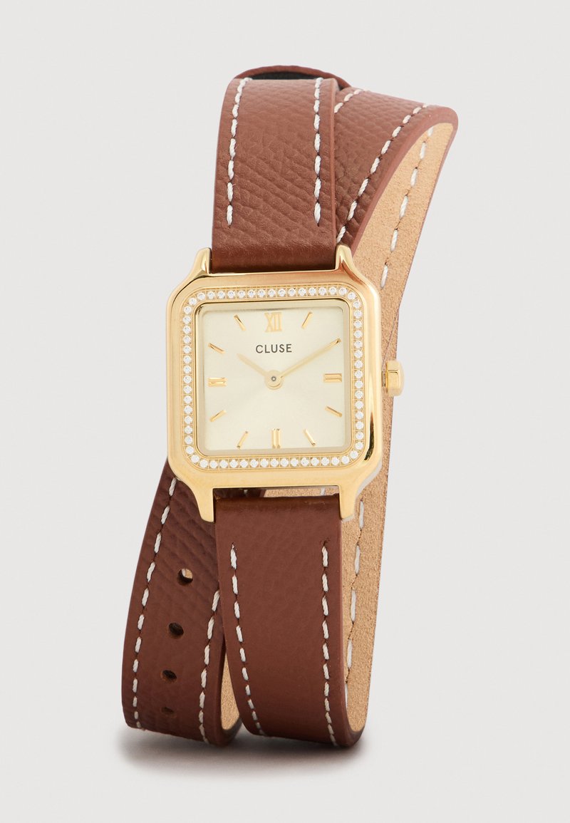 Zlaté štvorcové hodinky s krémovým ciferníkom, hnedým koženým remienkom s bielym prešívaním a obrubou posiatou kryštálmi. Značka: Cluse.