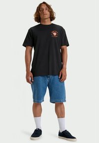 Hombre con cabello rizado, vestido con una camiseta gráfica negra, pantalones cortos de mezclilla azul, calcetines blancos y zapatillas negras, de pie contra un fondo liso.
