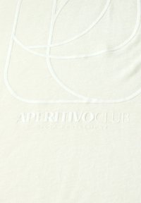 Vaaleanbeige T-paita, joka on valmistettu pehmeästä puuvillasta ja jossa on valkoiset ommellut viivat, jotka muodostavat abstrakteja muotoja sekä kohotettu logoteksti "APERITIVO CLUB".