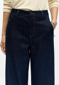 Tmavě modré džíny s vysokým pasem, viditelnými švy, předními kapsami a zapínáním na knoflík v pase. Hladká textura.