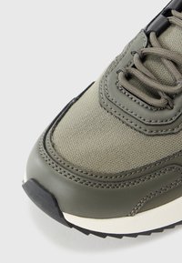 Olive green sneaker with textile upper, synthetic leather accents, rounded toe, textured sole, and contrasting cream midsole.