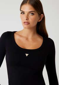Woman with straight brown hair wearing a black long-sleeve top with a square neckline and a small triangular logo at the center chest.