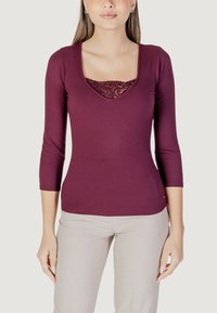 Maglietta a maniche lunghe bordeaux a coste con scollatura quadrata, caratterizzata da un inserto di pizzo sul petto. Abbinata a pantaloni chiari.