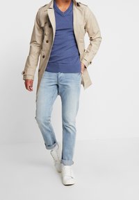Homme portant un trench-coat beige, un pull bleu à col en V, un jean bleu clair et des baskets blanches, marchant devant un fond blanc uni.