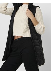 Eine Frau trägt eine schwarze, gesteppte ärmellose Jacke über einem cremefarbenen, gestrickten, kurzen Pullover und schwarzen Hosen, während sie den Kragen der Jacke hält.