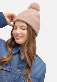 Gorro de punto rosa claro con pompón en la parte superior, con diseño acanalado y textura suave, usado por una persona con una camisa de mezclilla.