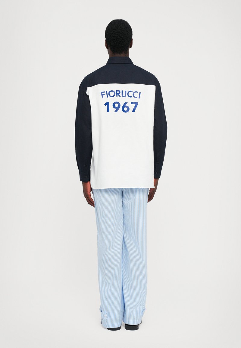 Fekete-fehér hosszú ujjú pólót viselő személy, melynek hátán "FIORUCCI 1967" felirat található, világoskék csíkos, bőnadrággal párosítva.