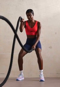 Red Nike sports bra with logo, navy shorts, white socks, and light blue athletic shoes. Engaged in battle rope exercise. Smooth background.
