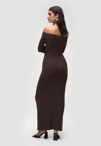 Abito maxi marrone con spalle scoperte, maniche lunghe, siluette aderente e una trama liscia. Presenta scarpe nere con tacco e punta quadrata.