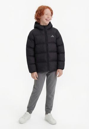 Niño pelirrojo de pie y sonriendo, vestido con una chaqueta acolchada negra, pantalones deportivos grises y zapatillas blancas sobre un fondo blanco.