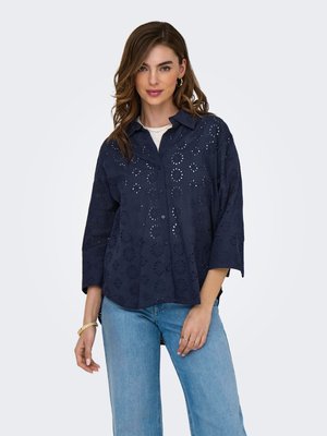 Kvinde med løst, bølget hår iført en mørkeblå skjorte med hulmønster over en hvid top og lyseblå jeans, stående mod en hvid baggrund.