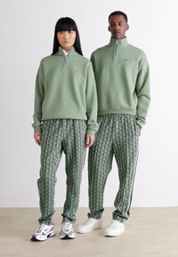 Two people stand side by side wearing matching light green zip-up sweaters and patterned green pants with white sneakers against a plain background.
