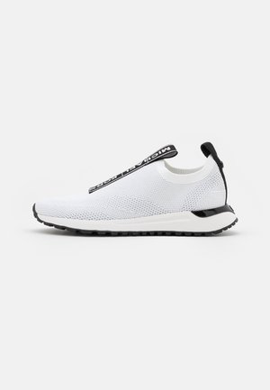 Hvid strikket slip-on sneaker med sort træktab og detaljer i sålen, med en sort rem mærket "MICHAEL KORS" over toppen.