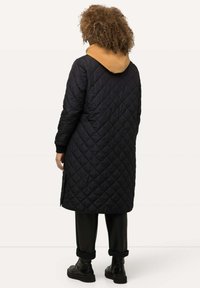 Quiltad svart lång jacka med en tan huva, med skräddarsydd passform och ribbade ärmslut. Svarta byxor och svarta stövlar fulländar outfiten.