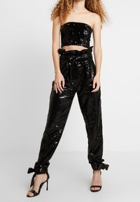 Černý korzetový crop top s flitry a odpovídající volné kalhoty s vysokým pasem a zavázaným pasem. Mezi doplňky patří manžetový lem a lesklá textura.