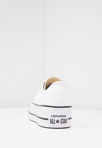 Baskets blanches à enfiler avec semelle en caoutchouc noir et bande. Présente une étiquette "Converse All Star" sur le talon. Texture lisse, design minimal.