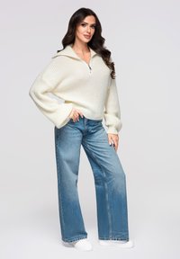 Pull-over en tricot blanc avec un col zippé, associé à un jean large bleu clair et des baskets blanches, présentant une silhouette décontractée.