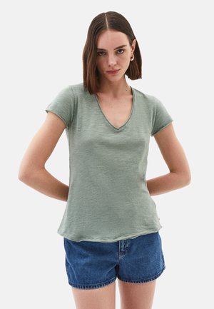 Grünes Kurzarm-V-Ausschnitt-T-Shirt aus strukturiertem Stoff. Es hat eine lockere Passform und einen abgerundeten Saum. Kombiniert mit blauen Jeansshorts.