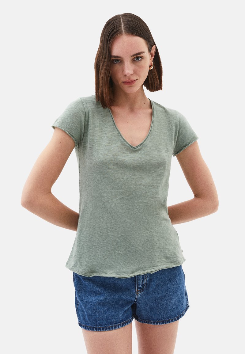 T-shirt verde a maniche corte con scollatura a V in un tessuto strutturato. Presenta una vestibilità comoda e un orlo curvato. Abbinato a pantaloni corti in denim blu.