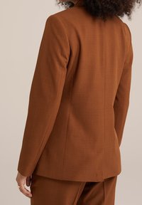 Brown tailored blazer with a smooth texture, single back vent, and subtle stitching details. Features a notched collar and structured shoulders.