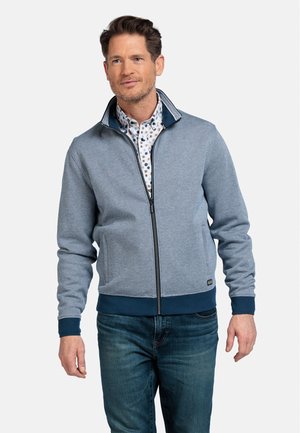 Man in een lichtblauwe gebreide zipjack over een witte met patroon shirt en blauwe spijkerbroek, staand tegen een effen witte achtergrond.