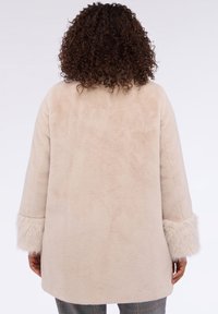 Beige tekoturkki, jossa on pehmeä rakenne, rento malli ja karvaiset hihansuut. Takakatsaus paljastaa sujuvan muotoilun ilman näkyviä kiinnityksiä.