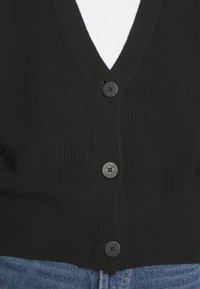 Gilet en tricot noir à col en V avec trois boutons noirs à motif, porté sur un haut blanc et un jean en denim bleu.