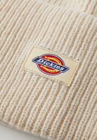 Krémová rebrovaná pletenina s prišitou béžovou nálepkou, na ktorej je logotyp značky Dickies v červenej, žltej, modrej a bielej farbe.