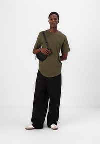 Tricou verde măsliniu cu mânecă scurtă, croială lejeră, asociat cu pantaloni negri largi și o geantă neagră tip crossbody. Pantofii au accente bej și albe.