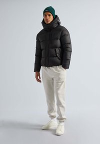 Schwarze Pufferjacke mit hohem Kragen und Reißverschlusstaschen, getragen über hellbeigen Jogginghosen und weißen Sneakers. Grüne Strickmütze auf dem Kopf.