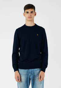 Maglione a maglia blu navy con colletto rotondo, dotato di un piccolo logo dorato sulla parte sinistra del petto. Indossato con jeans blu chiaro.