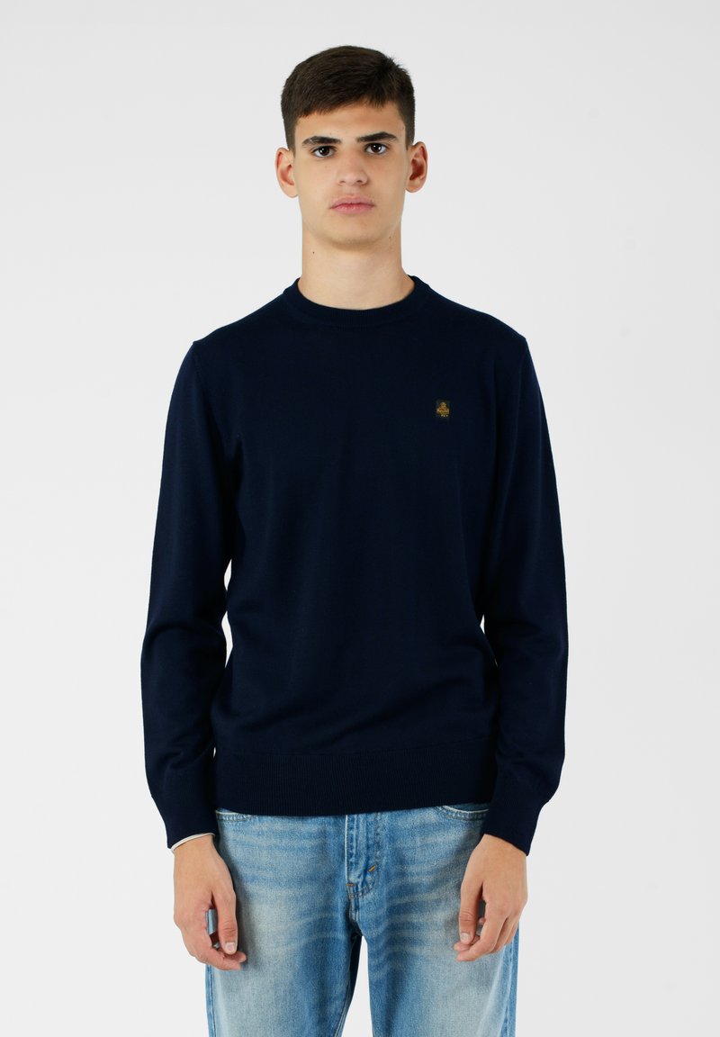 Maglione a maglia blu navy con colletto rotondo, dotato di un piccolo logo dorato sulla parte sinistra del petto. Indossato con jeans blu chiaro.