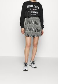 Zwarte sweatshirt met witte tekst, gecombineerd met een zwart-wit gestreepte rok in zigzagpatroon. Witte sneakers met zwarte accenten maken de look compleet.