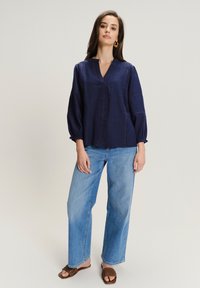 Granatowa bluzka z długim rękawem, z dekoltem w serek i haftowanymi detalami, w połączeniu z jasnoniebieskimi spodniami typu wide-leg i brązowymi sandałami na koturnie.