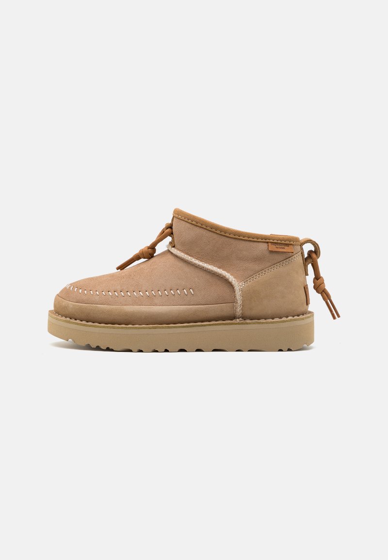 Beige suede ankle boots with a rounded toe, decorative stitching, and a tied accent on the side. Rubber sole with a textured grip.