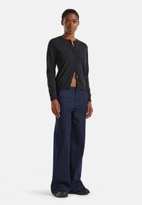 Cardigan noir à boutons avec un col rond, conçu dans un tissu tricoté doux ; associé à un pantalon en denim foncé à jambes larges et des baskets noires.