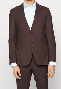 Costume en lin marron avec deux boutons, revers en dents de scie et poches latérales. Porté sur une chemise habillée bleu clair avec un col rabattable.