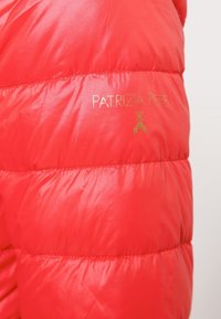 Punainen puffatakki, jossa on kiiltävä, tilkkutyylinen pinta. Kankaan pinnassa näkyy kultainen "PATRIIZIA PEPE" -logo ja yksityiskohtia.