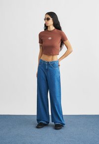 Καφέ κοντομάνικη T-shirt με λογότυπο, συνδυασμένη με φαρδιά μπλε τζιν παντελόνια. Αξεσουάρ περιλαμβάνουν γυαλιά ηλίου και μαύρα υποδήματα.