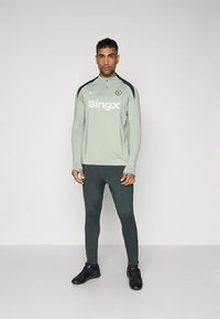 Roheline sportlik särk mustade aksentide ja logoga, mida on kombineeritud tumeroheliste keha vormivate pükstega ning mustade tennistega, mõeldud sporditegevuseks.