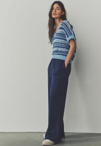 Pulover croșetat în tonuri de albastru, cu decolteu în V și mâneci largi, asortat cu pantaloni legați pe talie, de culoare bleumarin închis, cu croială largă.