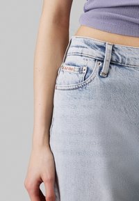 Jeans de mezclilla azul claro con una cintura alta, que presentan una pequeña etiqueta roja con el logo de Calvin Klein en el bolsillo y sutiles detalles de desgastado.