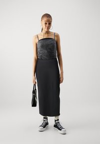Črn denim krojeni top z diskretnimi okraski, v kombinaciji z dolgo črno krilo. Model nosi črne visoke športne copate in drži črno torbico.
