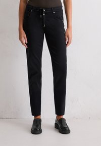 Persoon in zwarte slim-fit broek met koord in de taille en zwarte instap schoenen, staande tegen een lichte gestructureerde muur en vloer.
